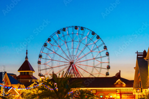 Evening amusement park with fabulous houses in garlands and a high Ferris wheel against the backdrop of a bright sky. Blurred image
