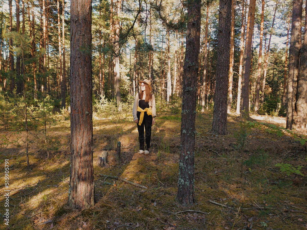 Fototapeta premium Young woman standing in the evergreen forest on a sunny summer day. Glasses, hat, denim overalls, sneakers. Mighty pine trees. Nature, hiking, trekking, ecotourism themes. Russia, Russian nature