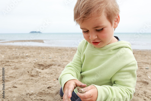 A little toddler 3years boy in yellow green hoodie is playing with sand on autumn spring beach cloudy day photo