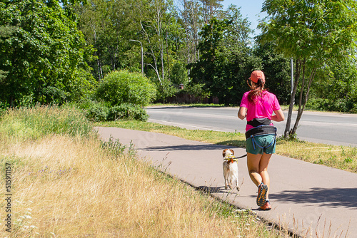 A female athlete in a pink T-shirt, shorts and a red baseball cap runs with a dog along the track. Canicross. Sports running with a dog. Lifestyle. photo