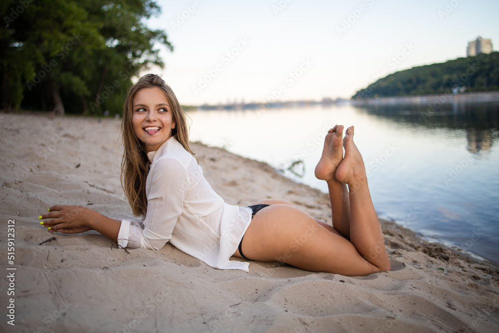 The beautiful sexy model in a white shirt posing against a setting sun near lake.