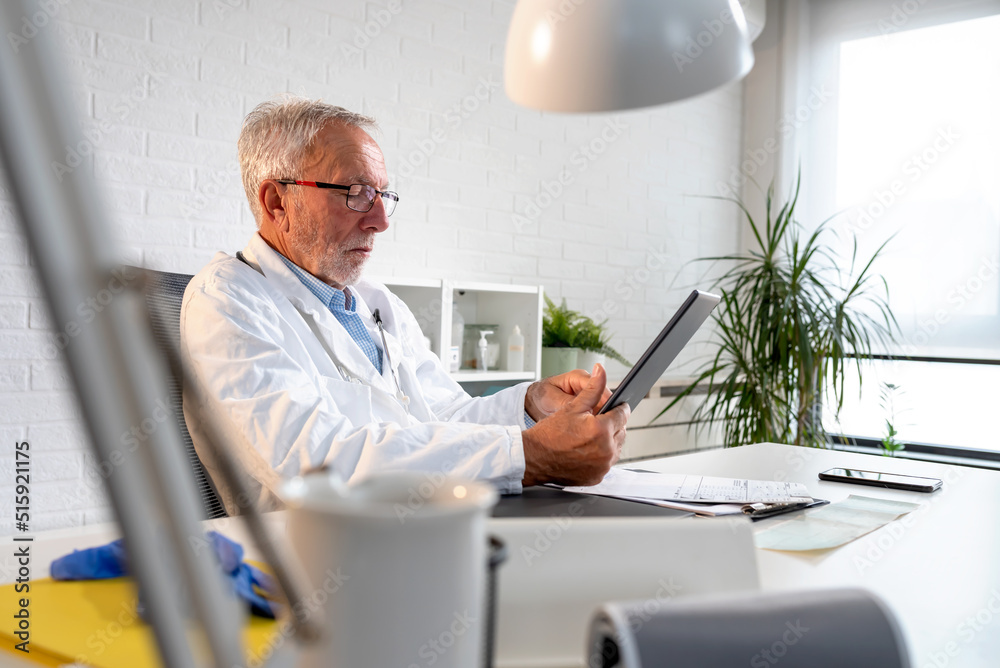 Experienced grey-haired doctor sitting in his office at the clinic using a tablet computer