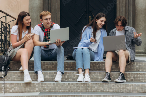 Group of cheerful students teenagers in casual outfits with note books and laptop are studying outdoors
