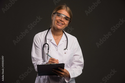 Blonde female doctor with a stethoscope wearing eyeglasses and writing a receipt to the patient while smiling
