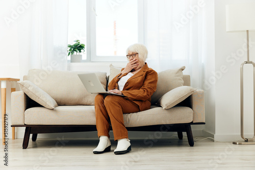 a shocked elderly woman with gray hair is sitting on a beige sofa in a brown suit working at a laptop and looking very emotionally amazed with surprise
