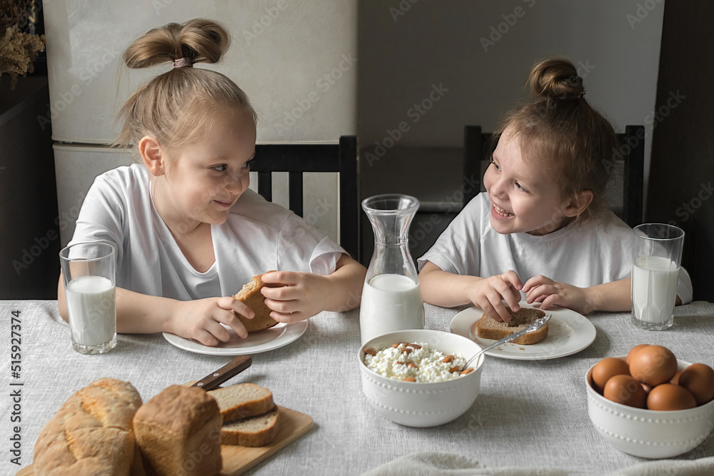 two cheerful sisters in white t-shirts have breakfast at home in the kitchen with natural and healthy products, cottage cheese and cornbread, milk and boiled chicken eggs