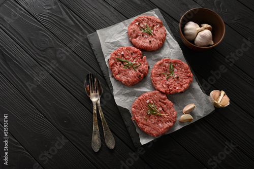 Raw beef burger patties on butcher's wooden board, rosemary and pepper. Black background. View from above.