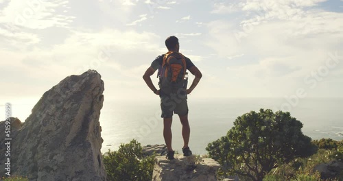 Rear view of fit active man standing with a backpack during a hike and enjoying the coastal landscape in front of him with seascape and coast. Male hiker standing on a rock looking at a nature view photo