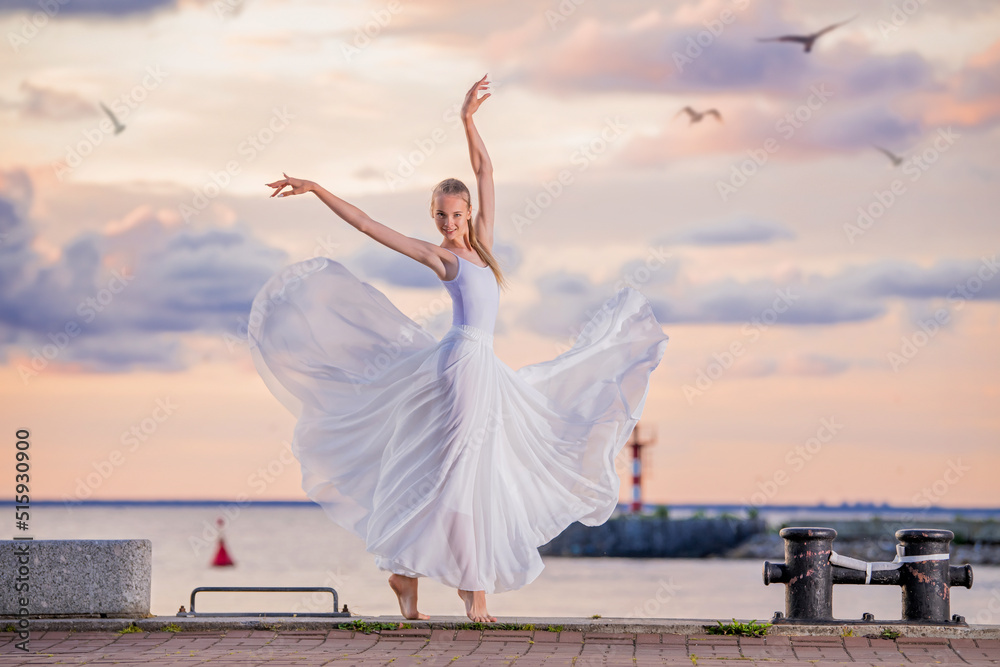 A dancing ballerina in a white flying skirt and tights on the ocean embankment or on the sea beach against backdrop of sunset sky A dancing ballerina in a white flying skirt and tights on the ocean embankment or on the sea beach against backdrop of sunset sky