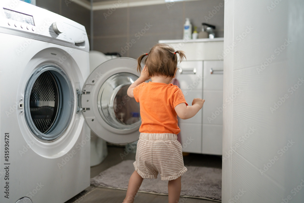 One girl small caucasian toddler child daughter standing at the washing machine in the toilet opening or closing the door examine and learn early development and growing up mischief concept copy space One girl small caucasian toddler child daughter standing at the washing machine in the toilet opening or closing the door examine and learn early development and growing up mischief concept copy space