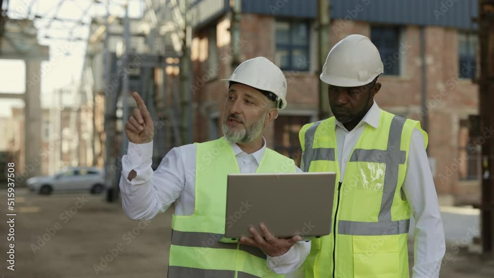 Caucasian aged architect holding portable laptop and explaining ideas to africa american construction worker. Multiethnic partners in protective uniform discussing structural process at building area. Caucasian aged architect holding portable laptop and explaining ideas to africa american construction worker. Multiethnic partners in protective uniform discussing structural process at building area.