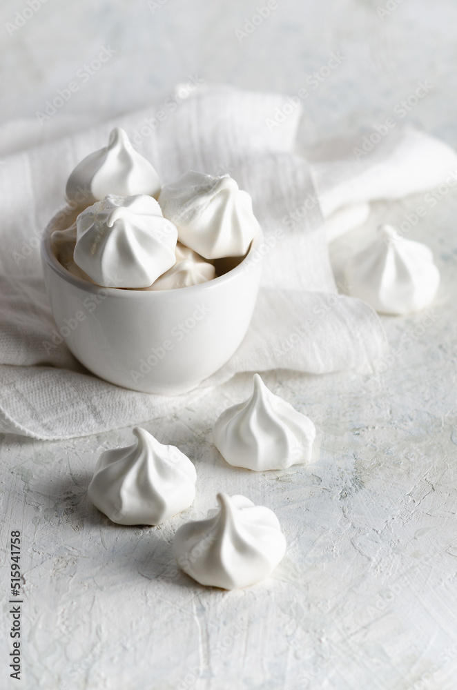 White meringue cookies in a white bowl with a white napkin, on grey background. White meringue cookies in a white bowl with a white napkin, on grey background.