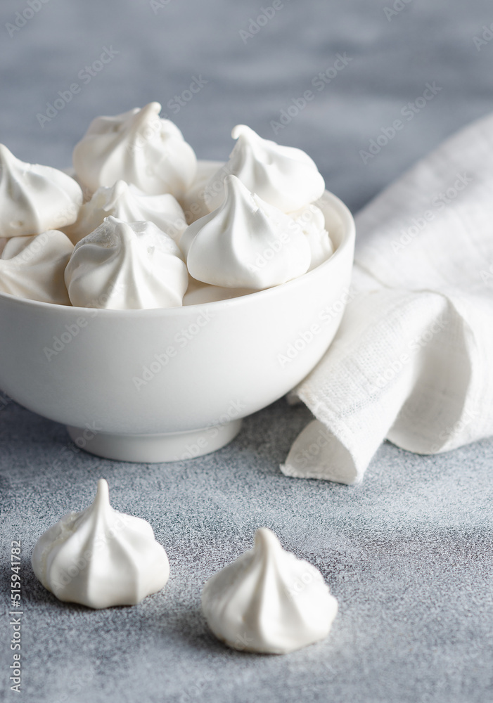 White meringue cookies in a white bowl with a white napkin, on grey background. 
