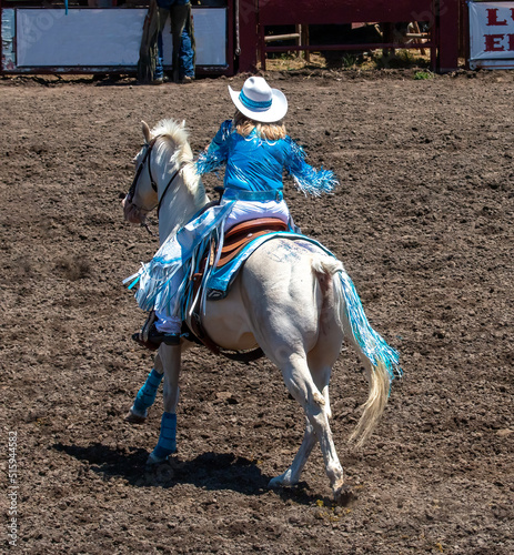 A rodeo cowgirl is part of a drill team. She is wearing blue fancy clothes and has a white hat. The horse is white with fancy sequences on its back. The arena is dirt.