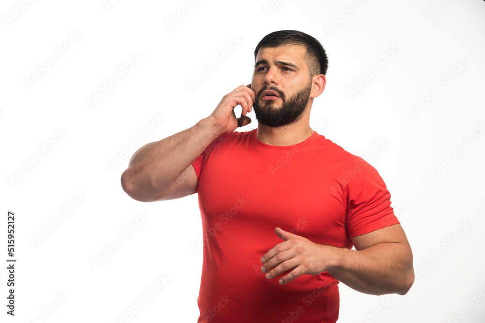 Sportive man in red shirt talking to the phone and demonstrating his arm muscles Sportive man in red shirt talking to the phone and demonstrating his arm muscles
