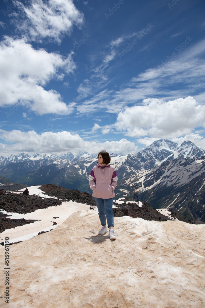 Cute teen girl in a pink jacket blue jeans and white sneakers standing on top of the mountains