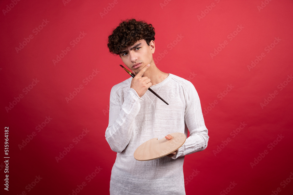 Young male artist posing with palette and brush on red background Young male artist posing with palette and brush on red background