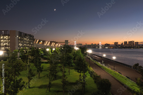 A wide shot of Toyosu Market Fisheries Intermediate Wholesale Market and Toyosu Gururi Park with the Rainbow Bridge, sunset Tokyo skyline, and the Moon in the starry sky.