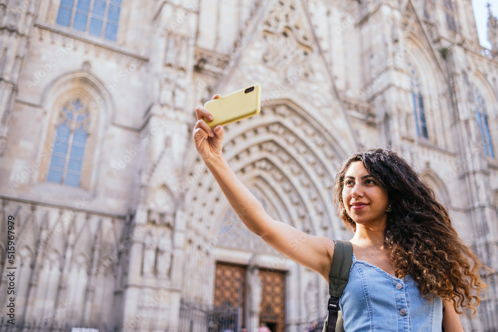 Low angle of young Hispanic woman with long curly hair taking selfie outside medieval cathedral on sunny summer day