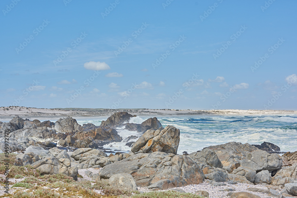 A shallow rocky coastline in the Cape Province, South Africa. Ocean waves crashing on coastal rocks and boulders on a sunny summer day, blue clear skies, and a scenic tropical landscape beachfront