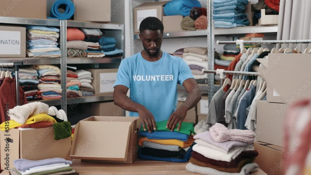 African american man standing at the warehouse while sorting and iterating clothes for donations. Belongings at the shelves at the background. Humanitarian aid concept African american man standing at the warehouse while sorting and iterating clothes for donations. Belongings at the shelves at the background. Humanitarian aid concept