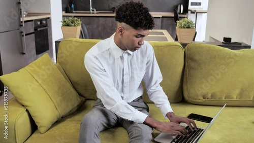 Young afro man freelancer using laptop device on cozy couch at home office, portrait of male working distantly on computer in modern interoir 4k photo