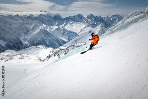 A skier in bright sports equipment goes down the glacier against the backdrop of a cloudy sky and snow-capped mountains
