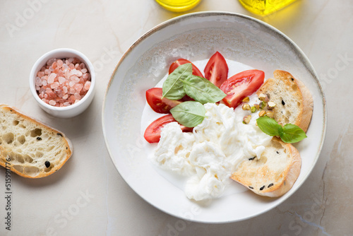 Plate of stracciatella or stretched curd cheese served with tomatoes, ciabatta and fresh basil, studio shot on a beige stone background