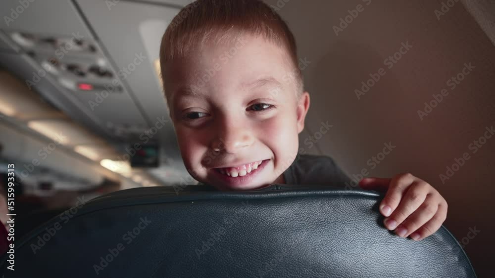 Child looks out between seats, leaning on chair and smiling. Flying in