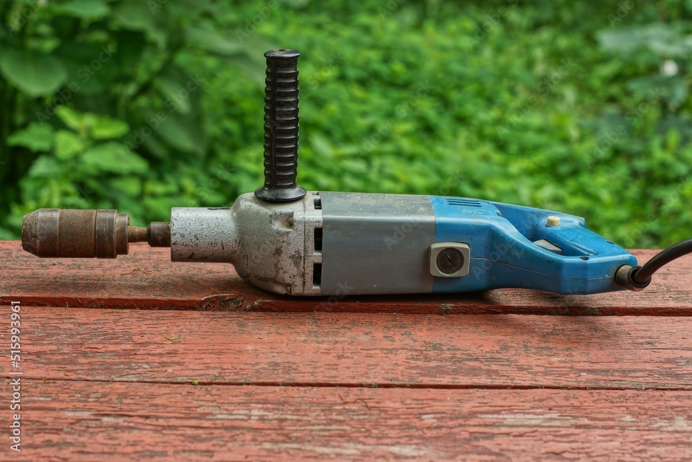 a very old powerful electric iron drill with a black handle and a rusty drill clamp lies on a wooden surface on a green background in the street during the day a very old powerful electric iron drill with a black handle and a rusty drill clamp lies on a wooden surface on a green background in the street during the day