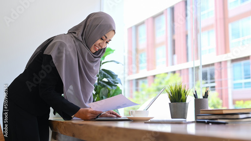 Professional young muslim business woman in hijab standing at office desk and checking financial data on her laptop computer