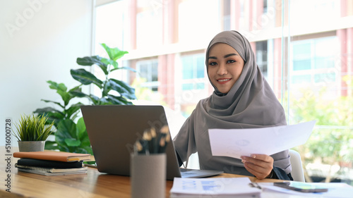 Young muslim businesswoman in hijab working with financial document and laptop computer at modern workplace