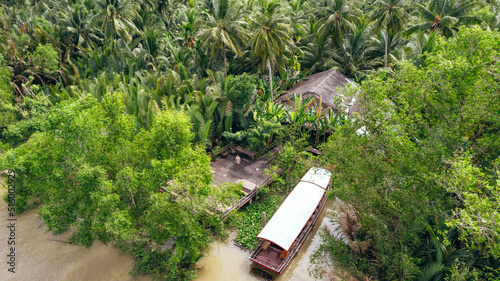 man standing on dock with boat on tropical coastline of Ben Tre along brown Mekong River with coconut tree jungle, aerial