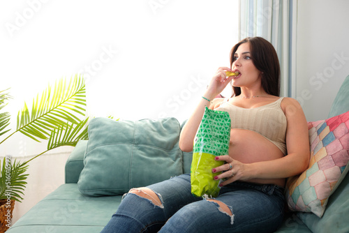 Pregnant young woman eating chips on the sofa at home. Food and cravings during pregnancy.