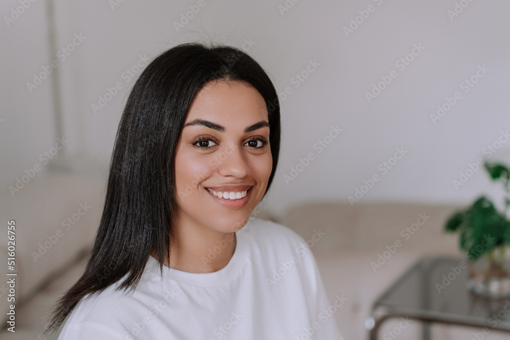 Portrait of African american gorgeous young woman with straight hair looking at camera toothy smiling at home in white t-shirt over blurry interior. Beauty and healthcare concept. Education and youth Portrait of African american gorgeous young woman with straight hair looking at camera toothy smiling at home in white t-shirt over blurry interior. Beauty and healthcare concept. Education and youth