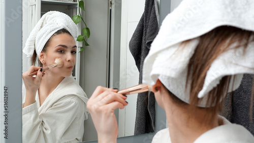 Young brunette woman applying foundation powder to her face with a makeup brush after showering. Bathroom morning routine in progress.