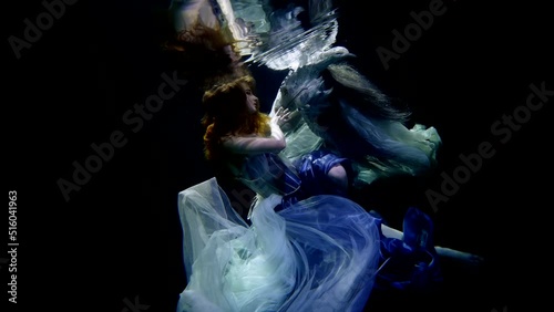 A young girl underwater in the contrasting lighting of the studio by the pool. The smooth fabric of her dress in weightlessness is like in a dream with a dragon.