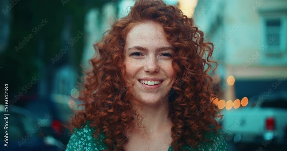 Portrait face young sunshine young woman with red hair look at camera smile stand in the city streets. Beautiful town at sunlight. Feel happy. Slow motion