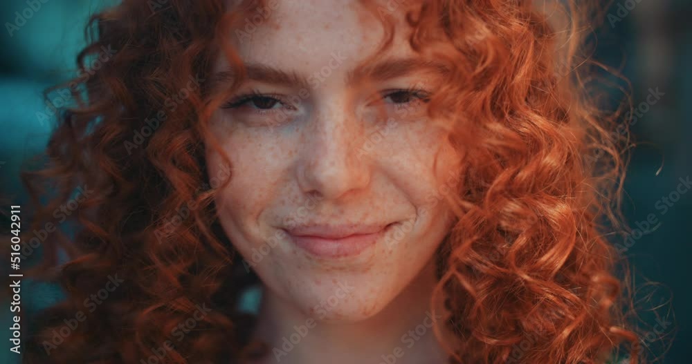 Close up face sunshine young smiling woman with red curly hair look at camera stand in the city streets. Feel happy. Summer. Outdoors at sunlight. Slow motion