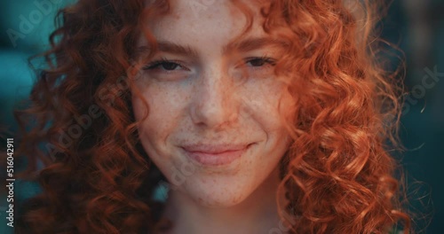 Close up face sunshine young smiling woman with red curly hair look at camera stand in the city streets. Feel happy. Summer. Outdoors at sunlight. Slow motion