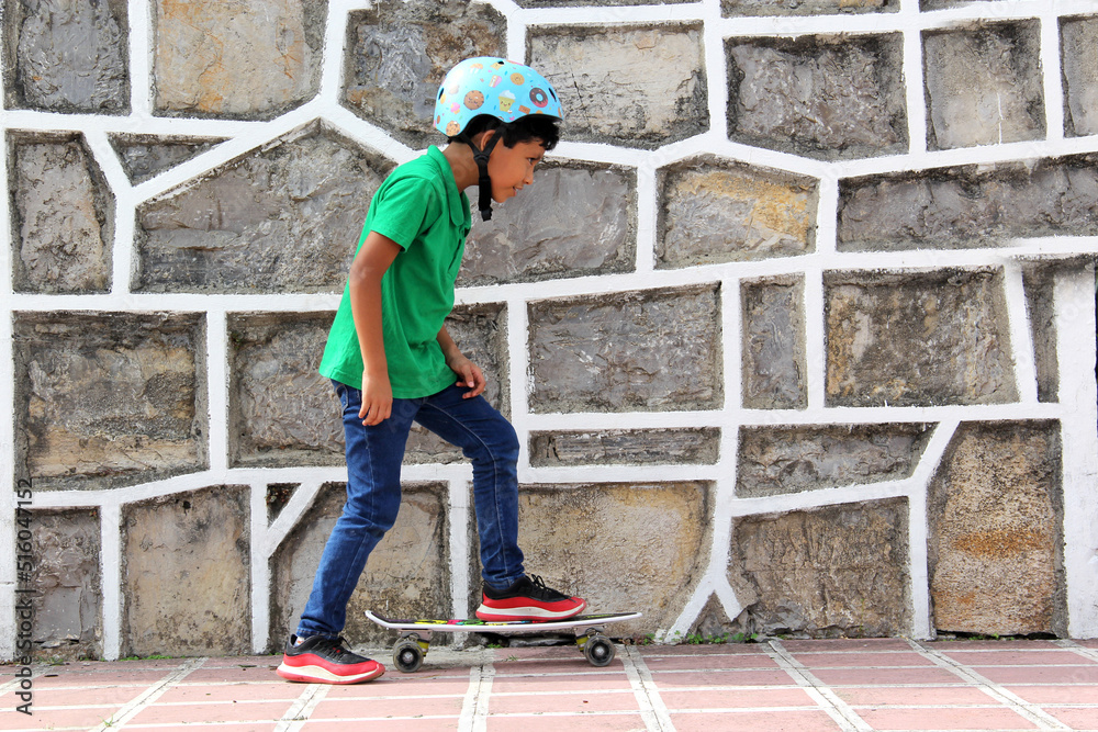 Little dark-haired Latino boy plays in the park with his skateboard wearing a helmet 