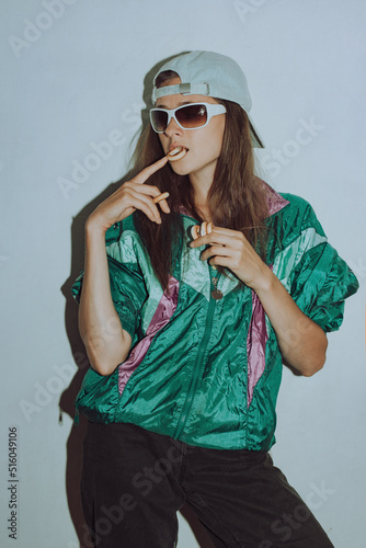 Cool teenager. Fashion girl in colorful trendy jacket and vintage retro sunglasses eating bagels in 80s - 90s style. Teenage girl in a cap on a white background.