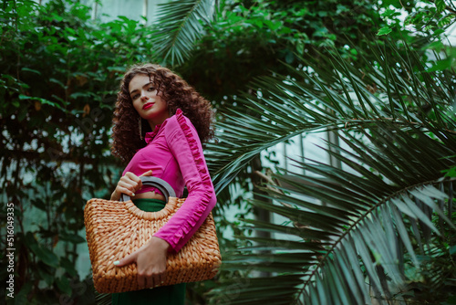 Fashionable curly woman wearing pink asymmetric ruffled top,  holding straw wicker top handle bag, posing in tropical garden. Summer fashion, lifestyle, beauty conception. Copy, empty space for text
