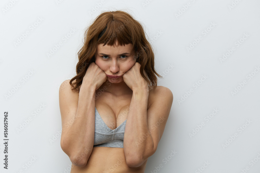 a funny, slightly upset tanned woman with red hair stands on a light background in a gray top and supports her cheeks with her fists, making a funny facial expression a funny, slightly upset tanned woman with red hair stands on a light background in a gray top and supports her cheeks with her fists, making a funny facial expression