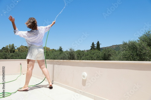 A mature woman in white clothes is cleaning a terrace of villa with water hose and dancing. Housewife has fun during houswork. photo