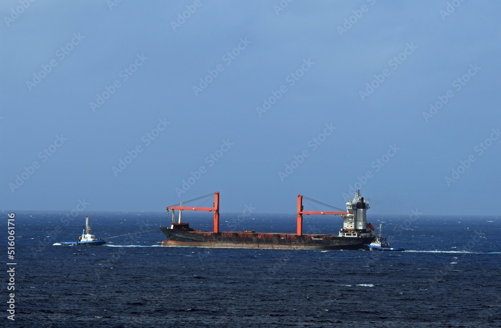 Large cargo ship with cranes being pulled by two blue and white tugboat, blue ocean with blue sky in the background
