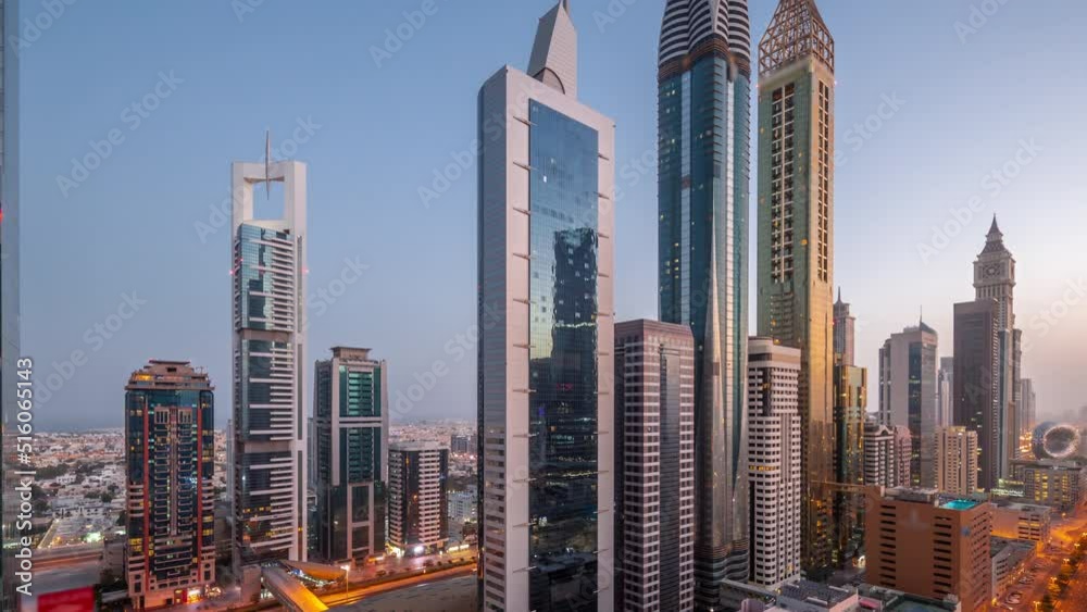 Aerial view of Dubai International Financial District with many skyscrapers night to day transition timelapse. Traffic on a road near metro station and multi storey parking with rooftop swimming pool