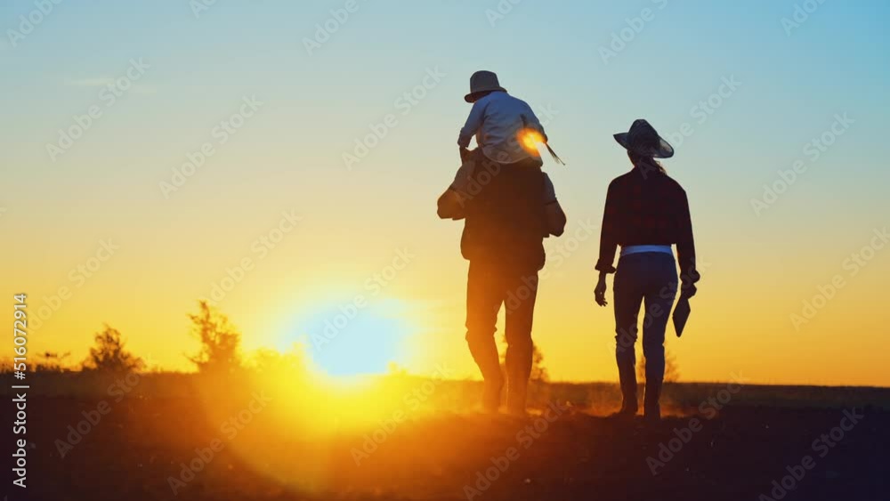Silhouette of family:child is sitting on dad shoulders and mom walking next on summer evening at sunset in field under summer sun. Boy holds hands plucked wildflowers ,farmer holds digital tablet. Silhouette of family:child is sitting on dad shoulders and mom walking next on summer evening at sunset in field under summer sun. Boy holds hands plucked wildflowers ,farmer holds digital tablet.