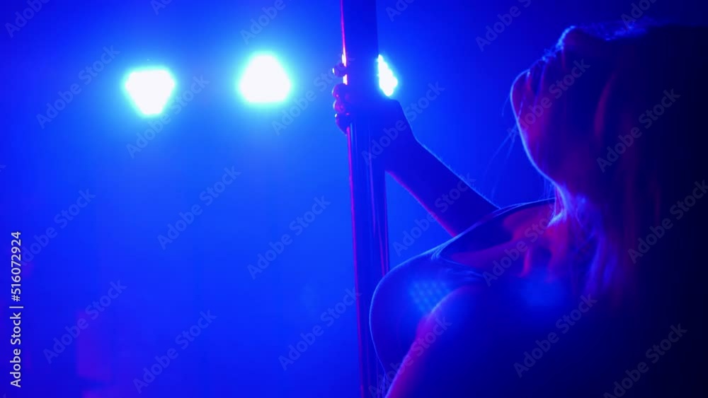 Close-up of a slender woman with long flowing hair near a pole in a nightclub, she performs a sensual dance in the dark under the light of multi-colored spotlights and smoke. Close-up of a slender woman with long flowing hair near a pole in a nightclub, she performs a sensual dance in the dark under the light of multi-colored spotlights and smoke.
