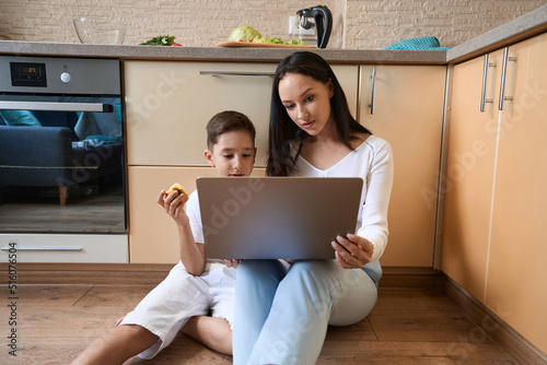 Boy studying online with his mother in kitchen